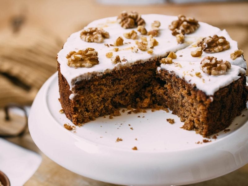 Ein angeschnittenes Stück Apfel-Walnuss-Kuchen auf einer weißen Etagere udn einem dunklen Holzbrett. Links daneben eine kleine Schale mit Walnüssen.