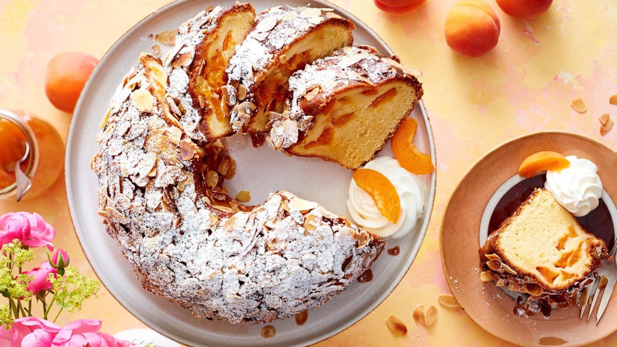 Ein angeschnittener Gugelhupf auf einem hellgrauen Teller mit Sahne und Aprikosen auf einem orange/rosa Hintergrund. Rechts ein Teller mit einem Stück Kuchen und links rosarote Blumen und ein Glas Aprikosenkonfitüre.