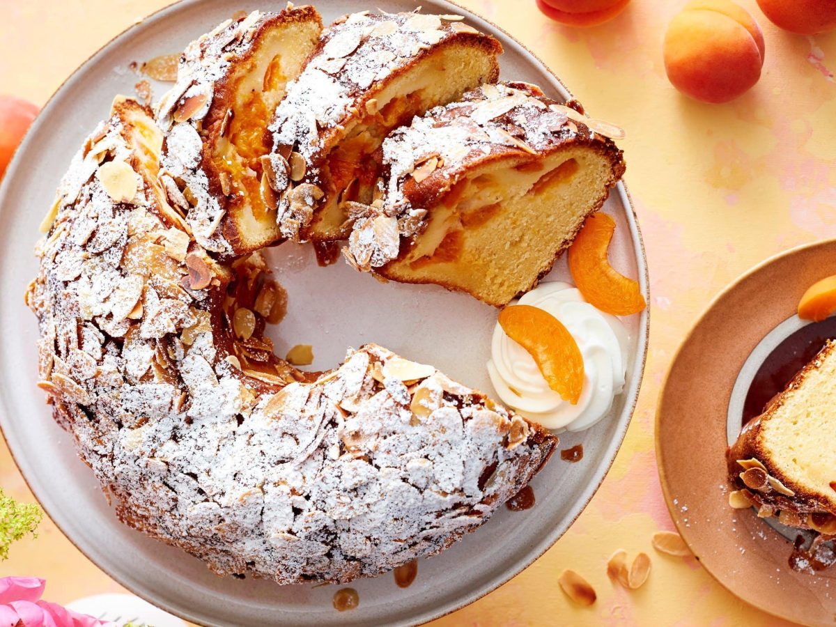 Ein angeschnittener Gugelhupf auf einem hellgrauen Teller mit Sahne und Aprikosen auf einem orange/rosa Hintergrund. Rechts ein Teller mit einem Stück Kuchen und links rosarote Blumen und ein Glas Aprikosenkonfitüre.