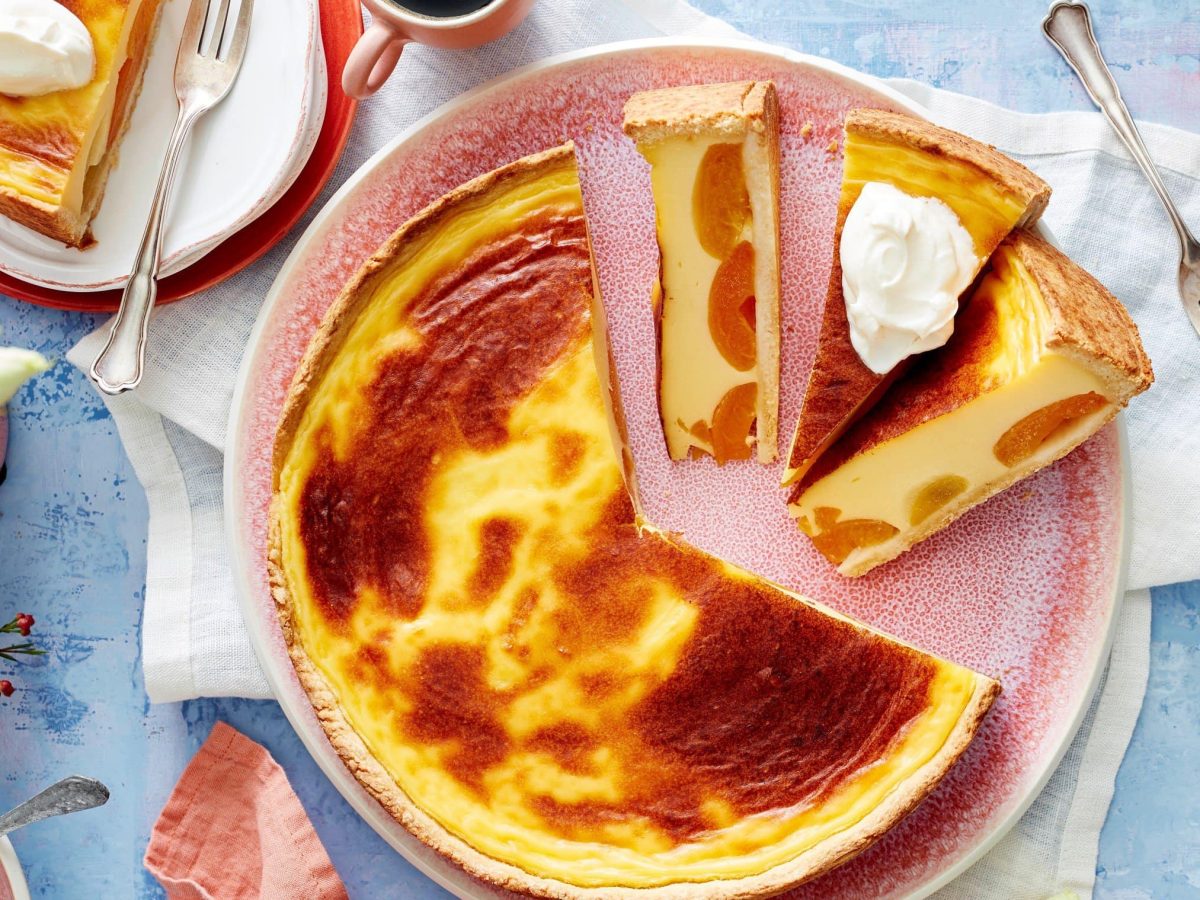 Angeschnittener Aprikosen-Puddingkuchen mit Schlagsahne auf einem rosa Teller. Darum auf blauem Untergrund Geschirr und eine Pflanze.