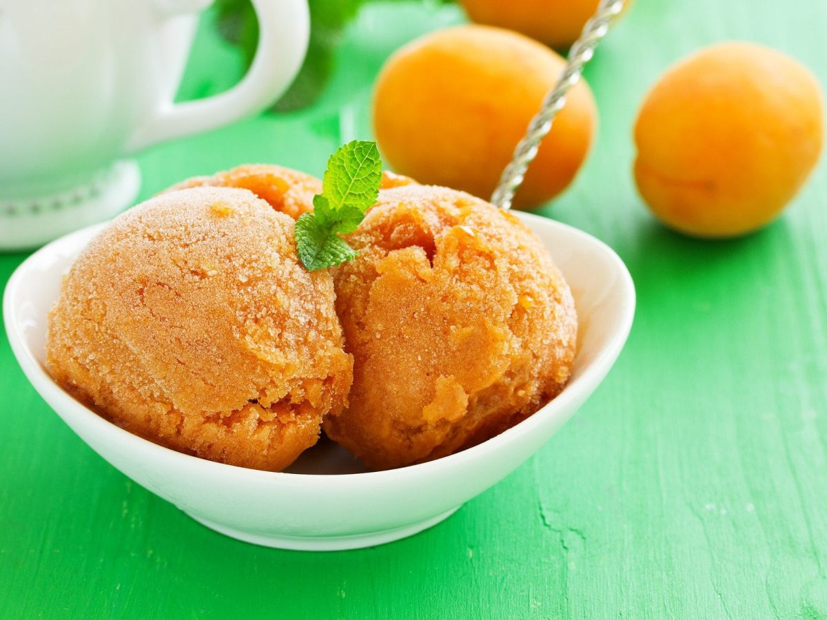 Drei Kugeln Aprikosen-Sorbet mit feiner Vanille ohne Eismaschine in weißer Schale mit Minzblatt auf grünem Untergrund. Im Bild eine weiße Kann und Aprikosen. Draufsicht.