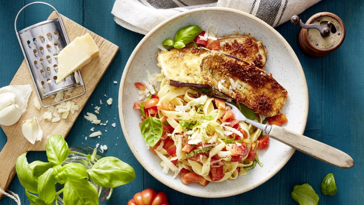 Ein Teller mit Auberginen-Schnitzel mit Bruschetta-Nudeln auf einem blauen Hintergrund. Links ein Schneidebrett mit Parmesan und einer Reibe. Links unten Basilikum in einem Glas.