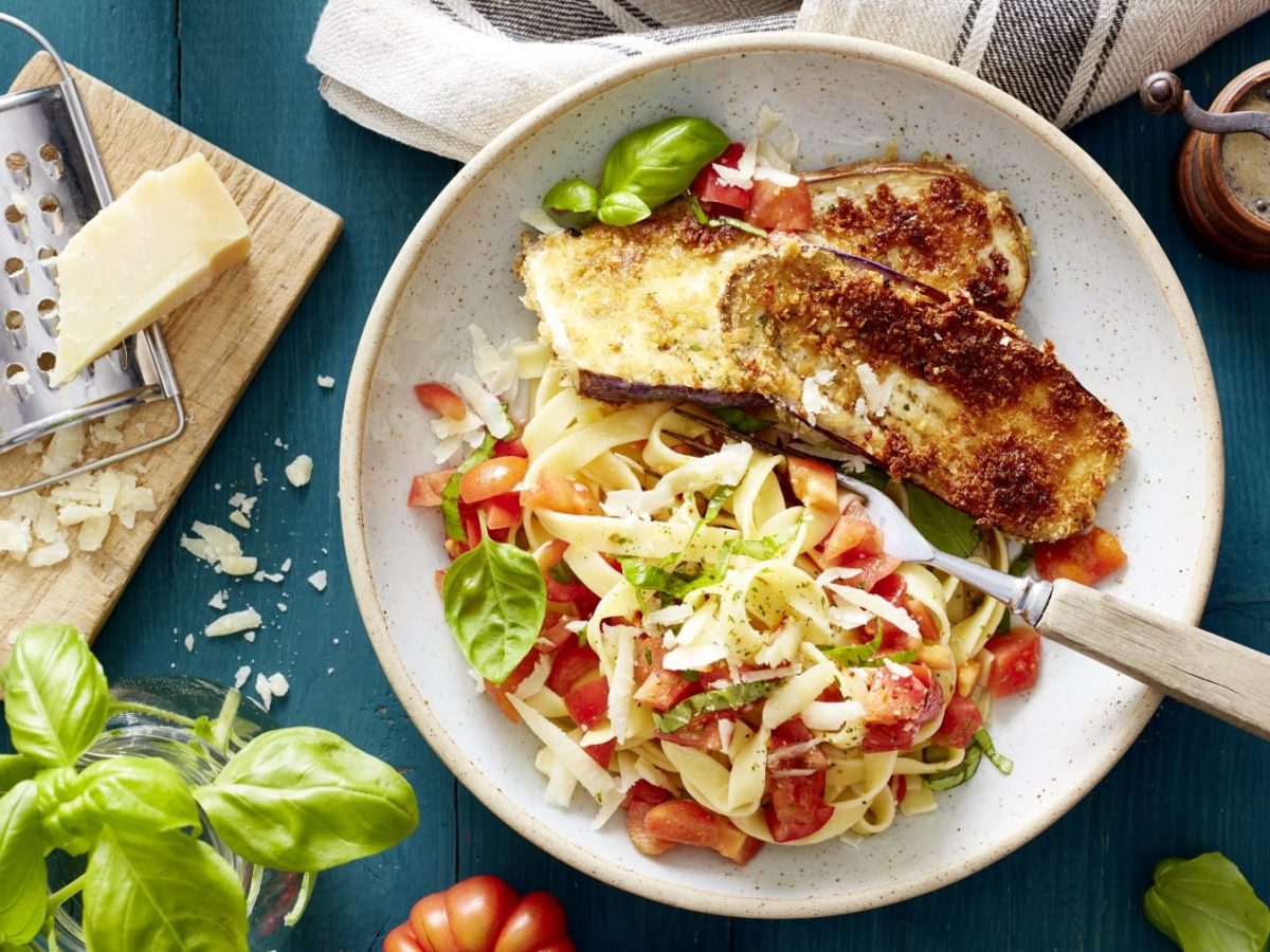 Ein Teller mit Auberginen-Schnitzel mit Bruschetta-Nudeln auf einem blauen Hintergrund. Links ein Schneidebrett mit Parmesan und einer Reibe. Links unten Basilikum in einem Glas.