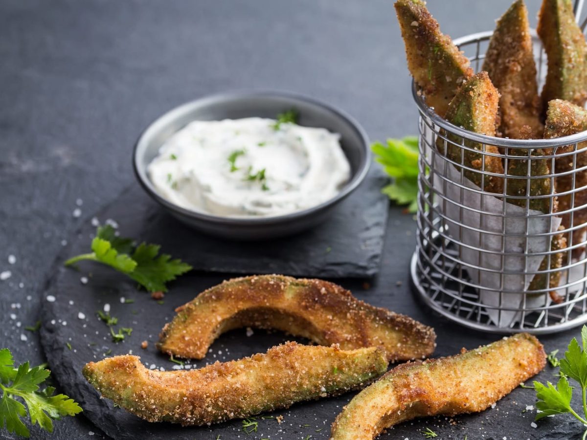 Drei Avocado-Fries auf einer runden Schiefertafel mit einem runden Frittierkorb aus Metall voller Avocado-Fries. Außerdem eine kleine Schale mit der weißen Sauce.