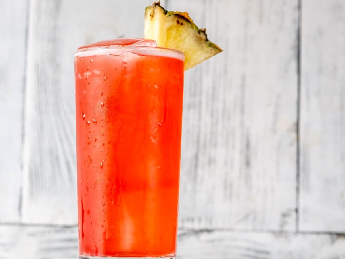 Brasilianischer Batida Rosa mit Cachaça, Grenadine und Ananas mit Ananasspalte im Highball-Glas vor hellem Hintergrund. Frontalansicht.