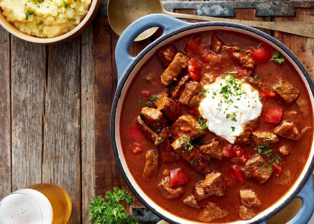 Ein Eintopf mit Gulasch auf einem Holztisch, darüber ein Suppenschöpfer. Links unten auf dem Tisch steht ein Bier und links oben ein Kartoffelpüree.