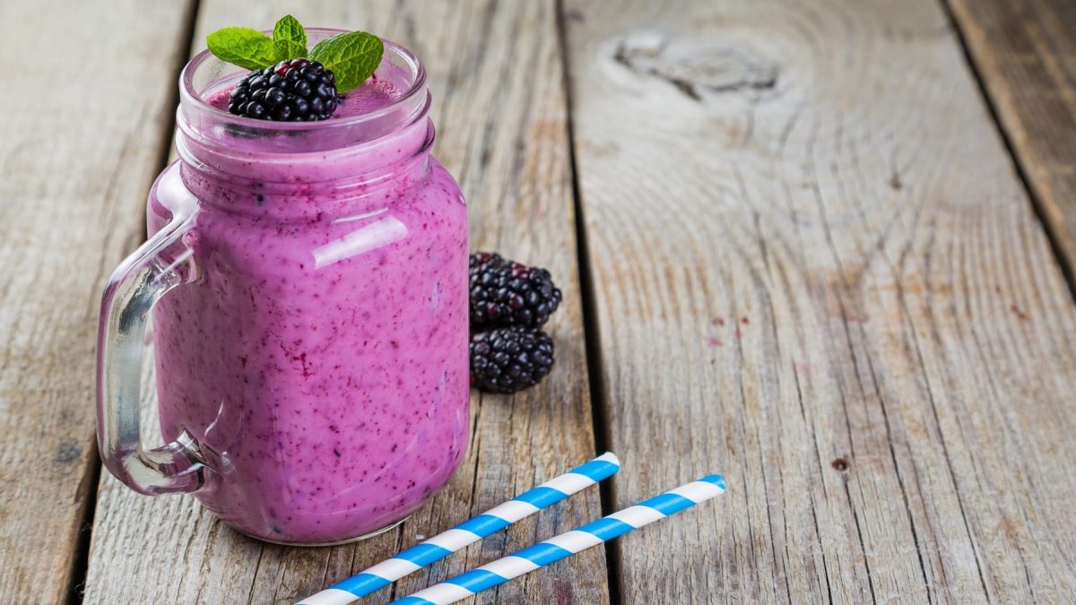 Ein Glas mit dem Brombeer-Joghurt-Smoothie auf einem Holztisch mit zwei blau-weiß-gestreiften Strohhalmen schräg von der Seite fotografiert.