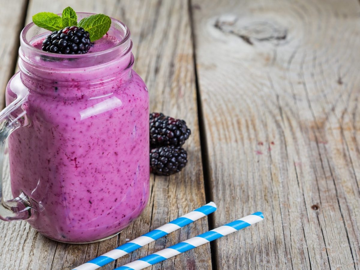 Ein Glas mit dem Brombeer-Joghurt-Smoothie auf einem Holztisch mit zwei blau-weiß-gestreiften Strohhalmen schräg von der Seite fotografiert.