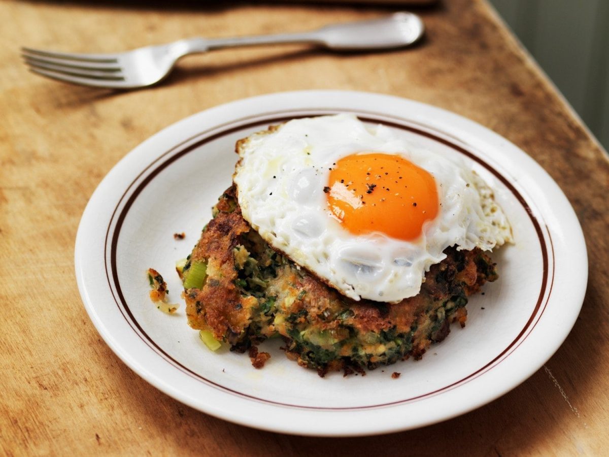 Draufsicht: Auf einem Holztisch steht eine weißer Runder Teller mit Bubble and Squeak mit einem Spiegelei. Daneben liegt eine Gabel.