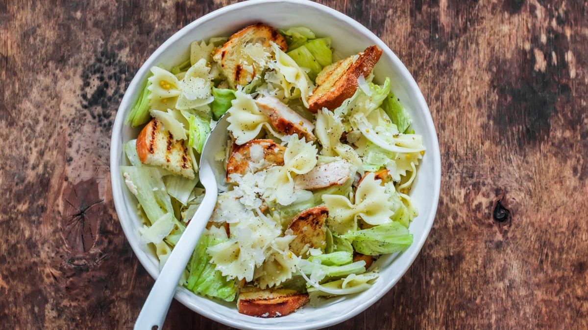 Eine weiße Schüssel auf einem Holzuntergrund mit einem Caesar-Salad mit Nudeln.