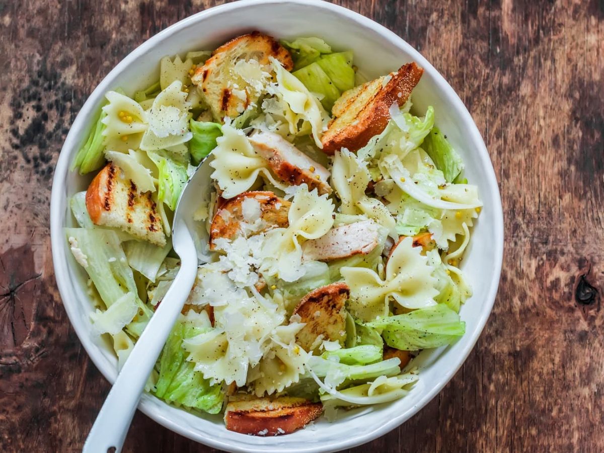 Eine weiße Schüssel auf einem Holzuntergrund mit einem Caesar-Salad mit Nudeln.