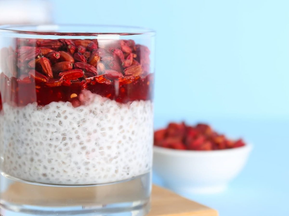 Ein kleines Glas mit dem Chia Pudding und darauf Himbeeren und Goji-Beeren auf der linken Seite des Bildes. Alles vor hellblauem Unter- und Hintergrund.