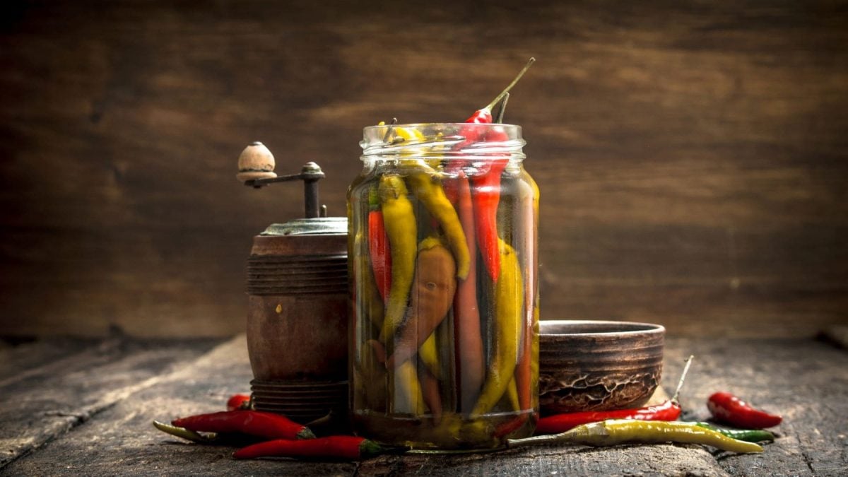 Selbst eingelegte Peperoni, Chilis oder Pfefferonen im offenen Glas mit Gewürzmühle auf Holz. Frontalaufnahme.