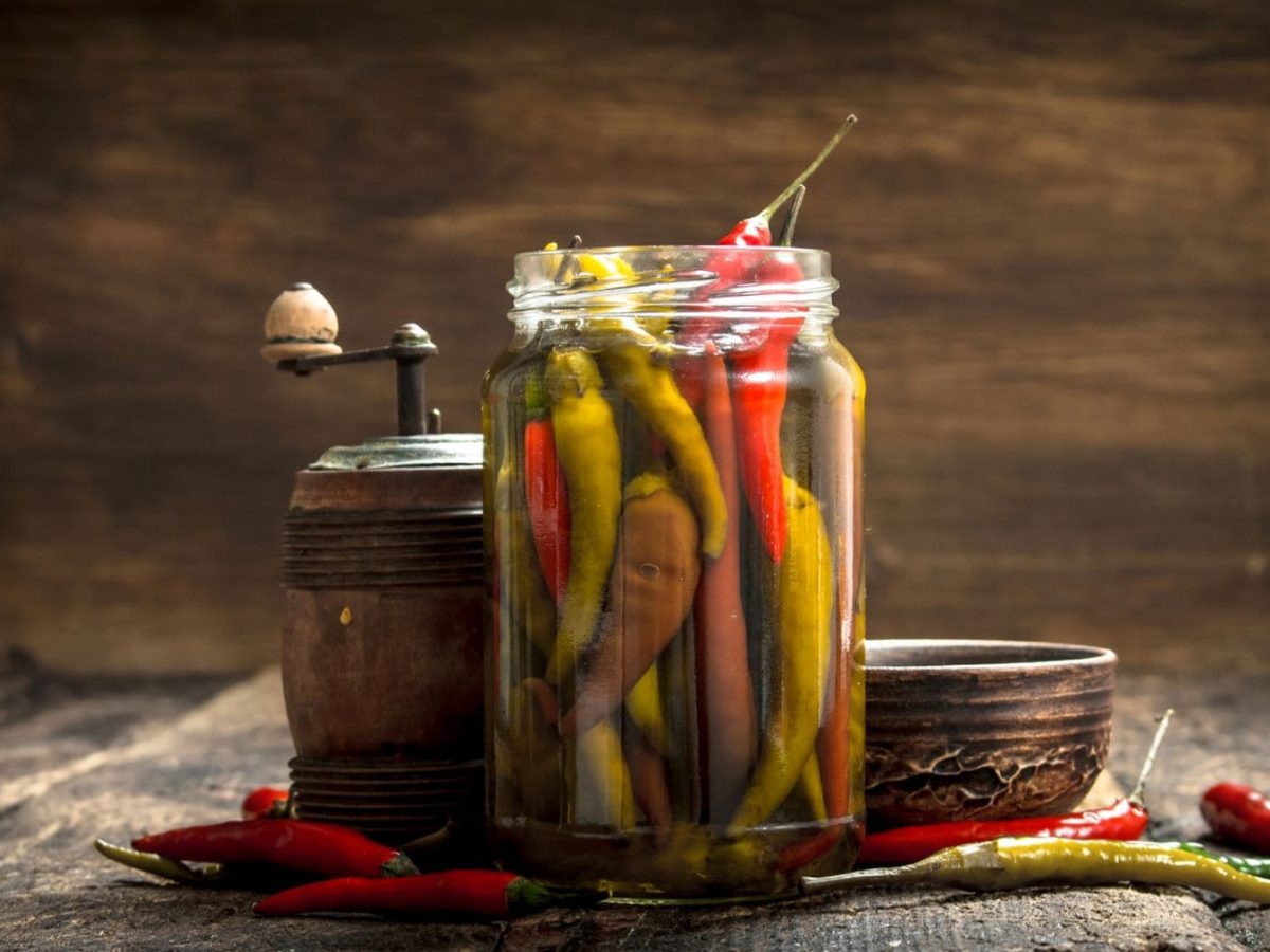 Selbst eingelegte Peperoni, Chilis oder Pfefferonen im offenen Glas mit Gewürzmühle auf Holz. Frontalaufnahme.