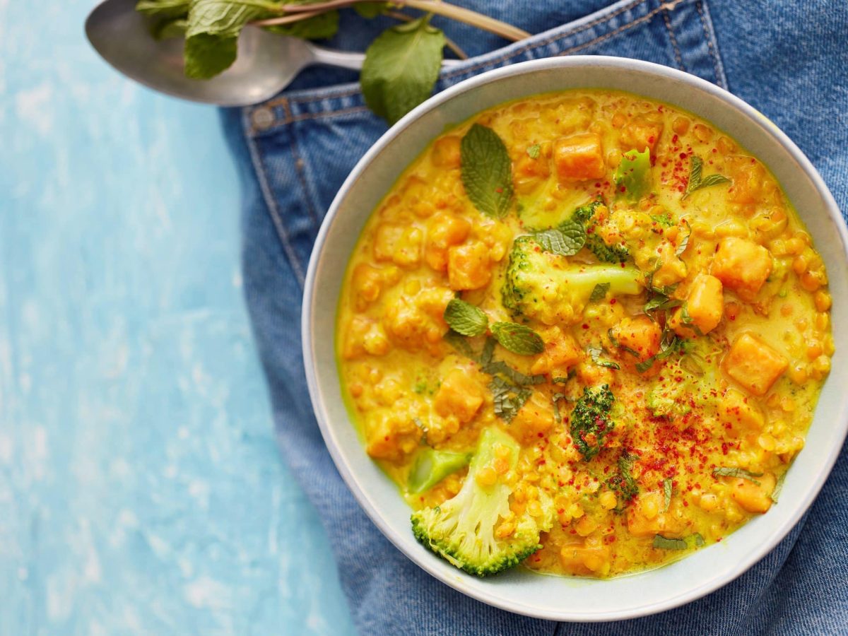 Eine hellgraue Bowl mit rotem Linsencurry auf einem Jeansstoff auf einem hellblauen Hintergrund. Oben ein Löffel und Minzblätter.