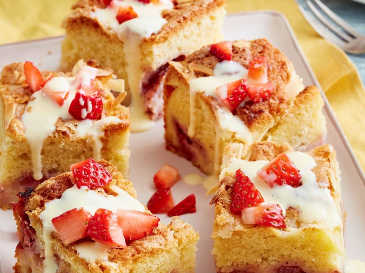 Angeschnittener Erdbeer-Pudding-Butterkuchen in Vierecken mit Guss und frischen Erdbeeren drauf auf einem beigen Teller auf Holzuntergrund.