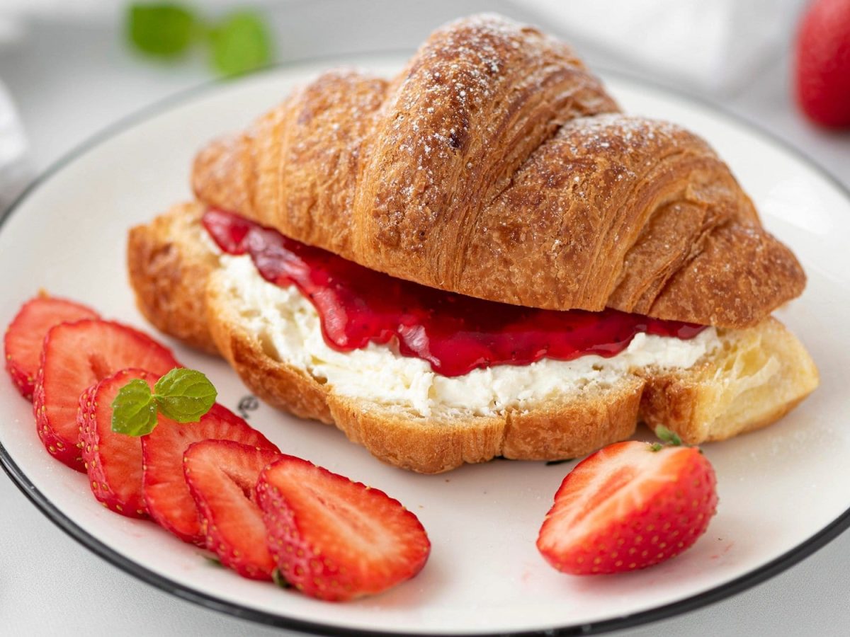 Auf einem weißen Teller liegt ein Erdbeer-Croissant mit Ricotta-Minze. Neben dem Gebäck liegen mehrere Erdbeerscheiben.