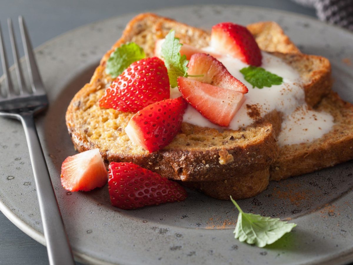 In Ahornsirup getränkter French Toast mit Erdbeeren und Vanillejoghurt auf Teller mit Gabel und Minze auf grauem Untergrund. Draufsicht.