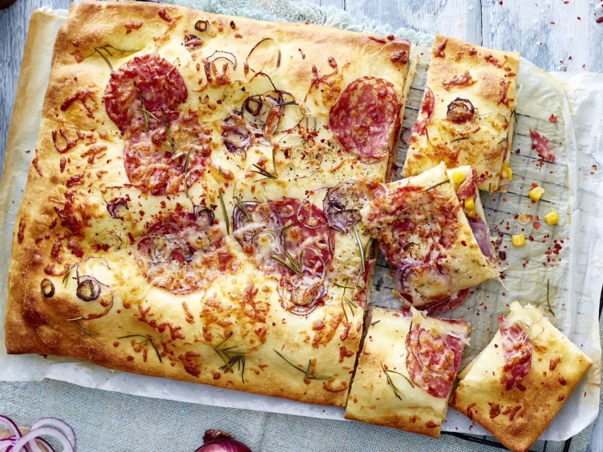 Ein Backofenrost mit Backpapier und gefüllter Pizza auf einem gräulichen Hintergrund. Links oben ein Bund Rosmarin, rechts oben eine kleine Bowl mit Chilisalz. Links unten ein Schneidebrett mit geschnittenen Zwiebeln und rechts unten ein Teller mit einem Stück Fugazetta.