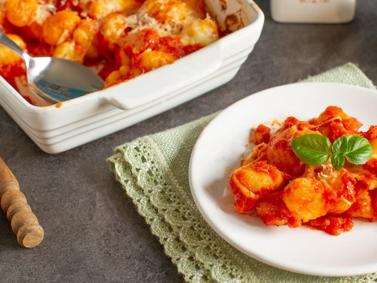 Ein weißer Teller mit dem Gnocchi-Ofengericht und der Auflaufform auf einem grauen Tisch mit Besteck und einer grünen Tischdecke.