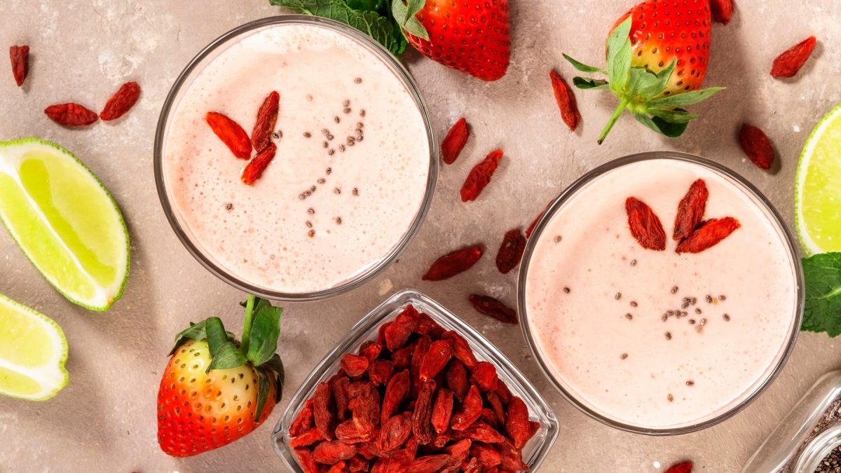 Zwei Gläser mit dem Shake von oben fotografiert. Daneben zahlreiche Zutaten des Shakes wie die Erdbeeren, Goji-Beeren und Limettenspalten.