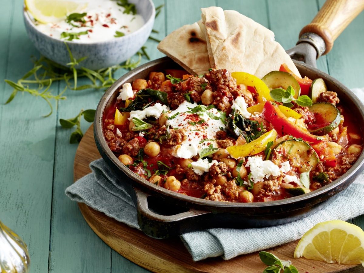 Eine Griechische Hack-Gemüse-Pfanne auf einem Geschirrtuch auf einem Holzbrett auf einem grünen Hintergrund. Links oben eine weiße Sauce in einer kleinen Schüssel, links unten eine Flasche mit Olivenöl.