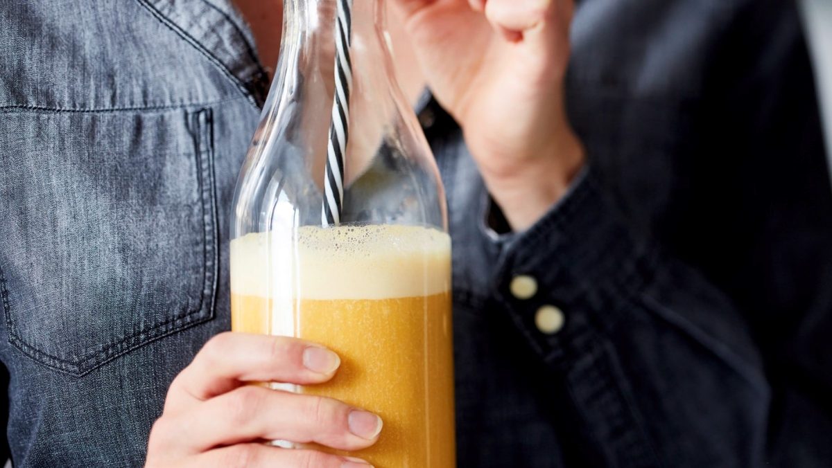 Eine Glasflasche gefüllt mit einem gelben Smoothie und einem schwarz-weiß gestreiften Strohhalm, gehalten von einer Person. Nur der Oberkörper ist sichtbar.