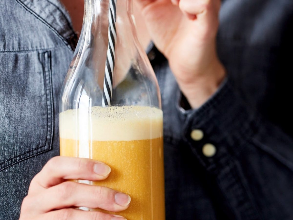 Eine Glasflasche gefüllt mit einem gelben Smoothie und einem schwarz-weiß gestreiften Strohhalm, gehalten von einer Person. Nur der Oberkörper ist sichtbar.