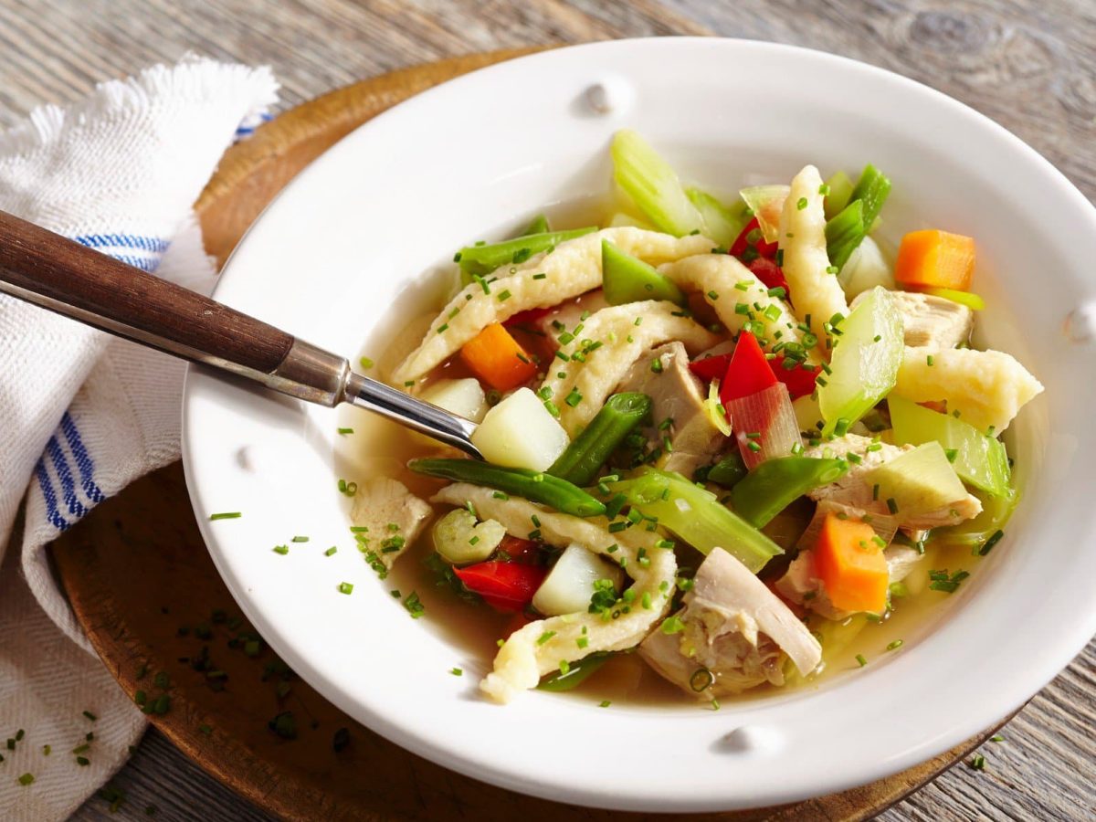 Hühnersuppe mit Pizokel und Sommergemüse und Schnittlauch in weißem Suppenteller auf Holztisch. In der Suppe ein Suppenlöffel mit Holzgriff. Links ein blau-weißes Küchentuch. Draufsicht.