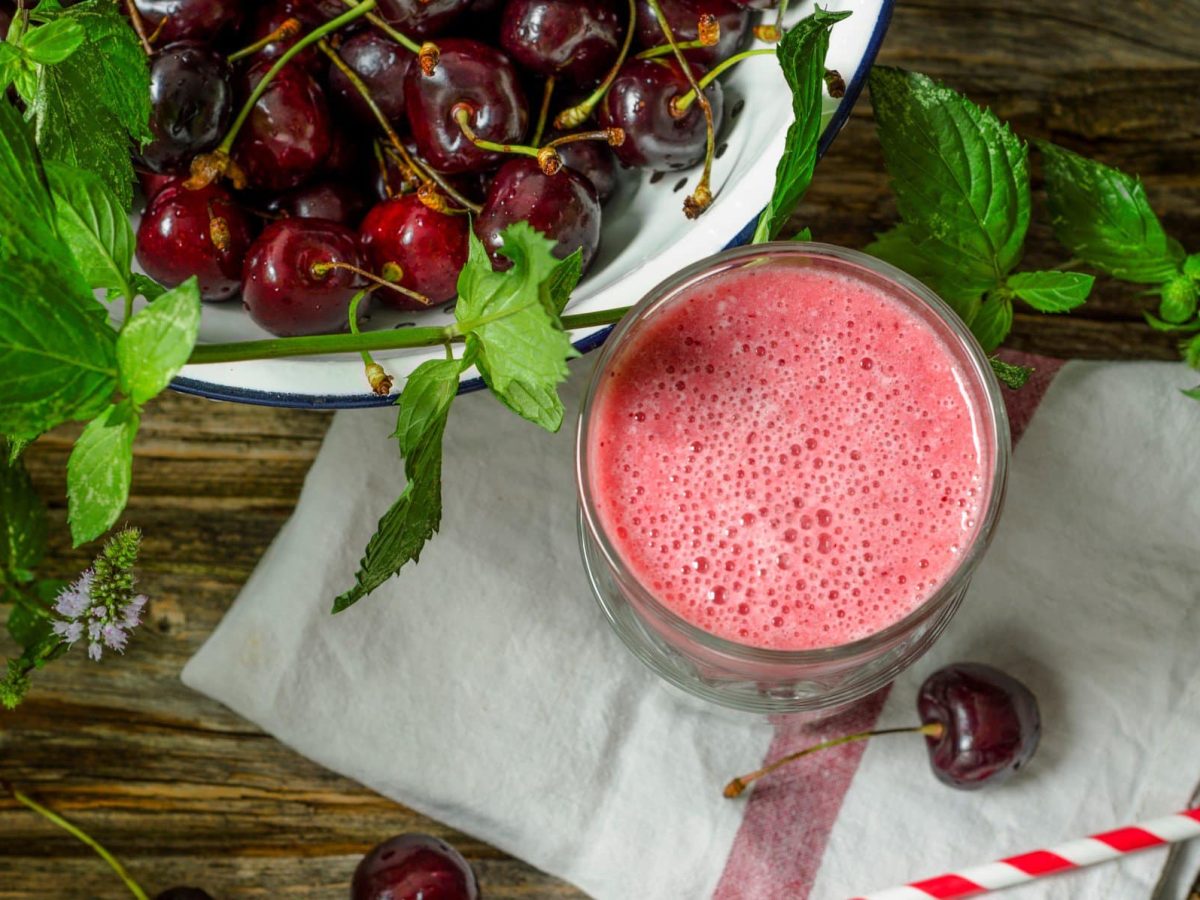 Kirsch-Smoothie mit Skyr und frischer Minze im Glas auf grauem Tuch und Holztisch. Daneben eine Schale mit Kirschen und Minzstängeln. Im Bild ein rot-weißer Strohhalm, eine Kirsche und Minzblätter. Draufsicht.