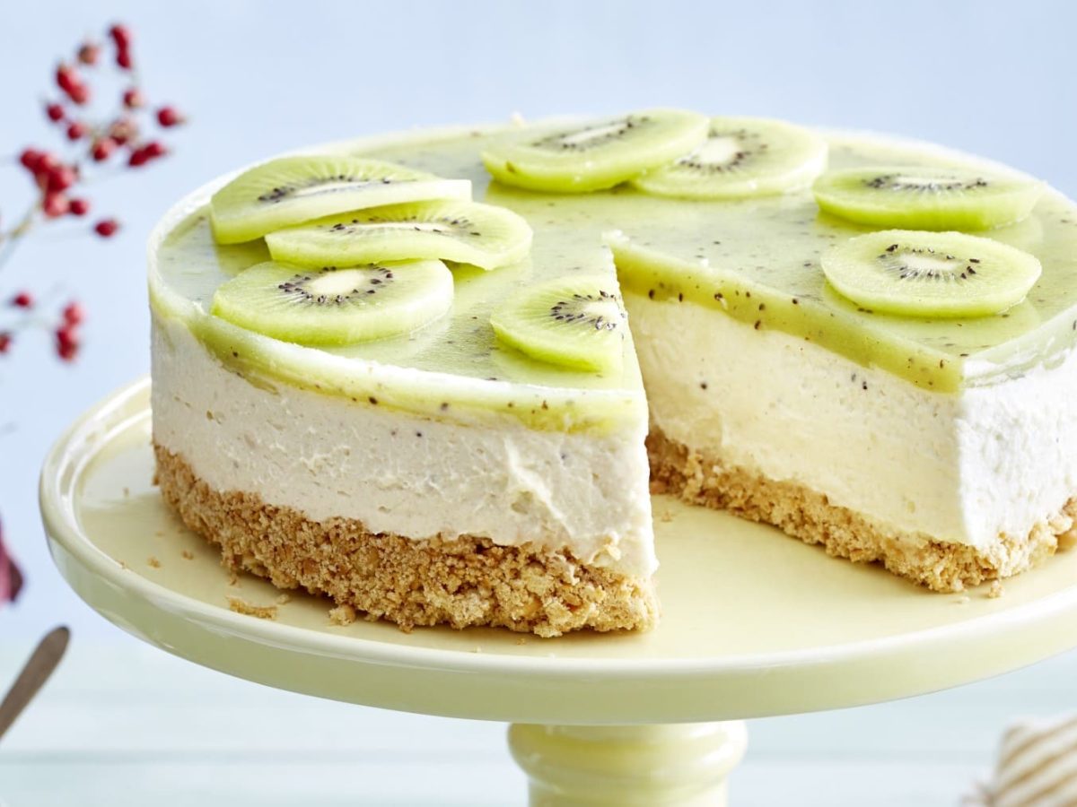 Ein Kiwi-Cheesecake auf einer hellgelben Tortenplatte vor einem bläulichen Hintergrund. Links ein Blumenstrauß.