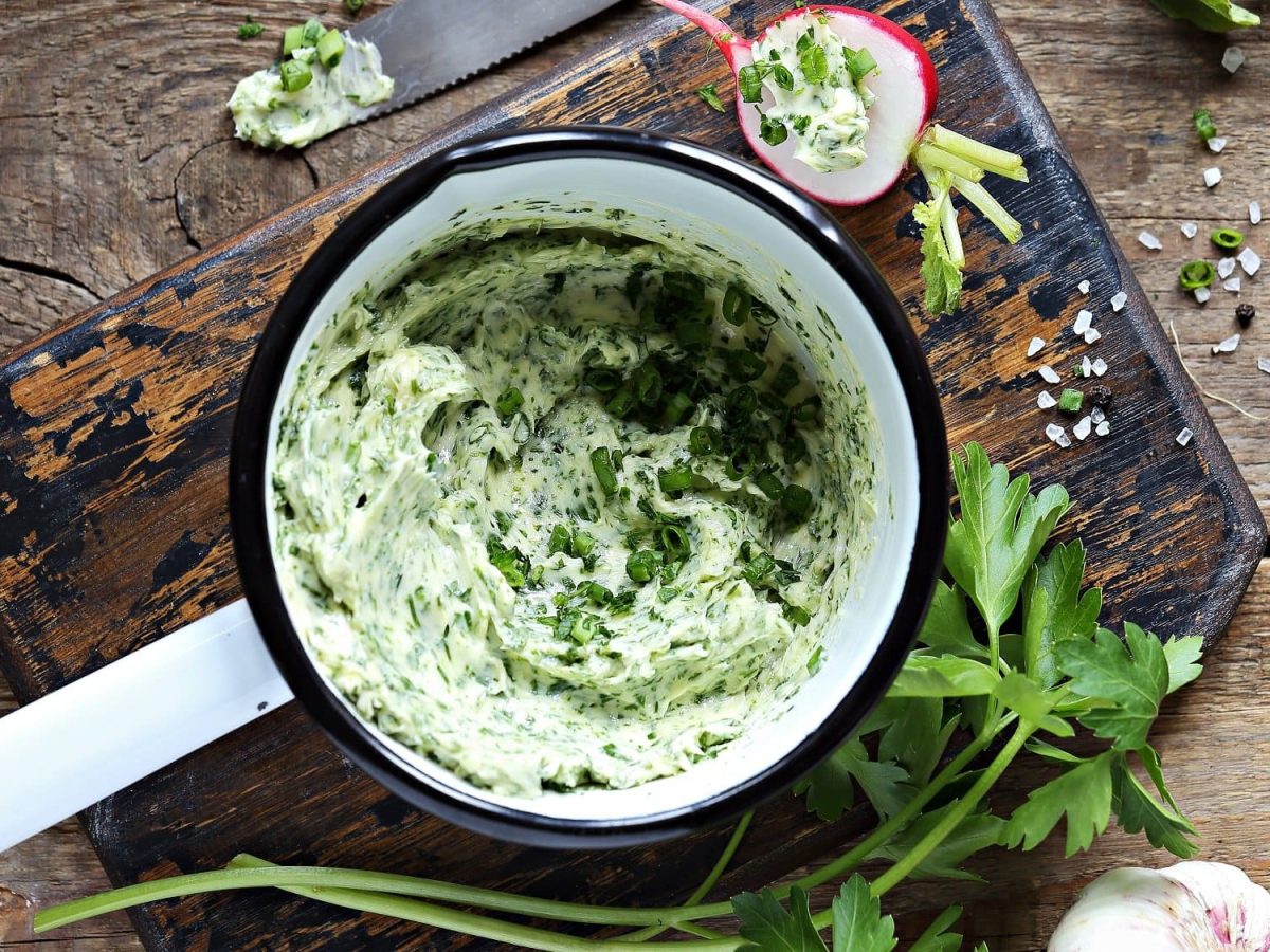 Eine Emaille-Schale mit Knoblauch-Parmesan-Butter in der Draufsicht, drumherum frische Petersilie, ein Messer und Radieschen.