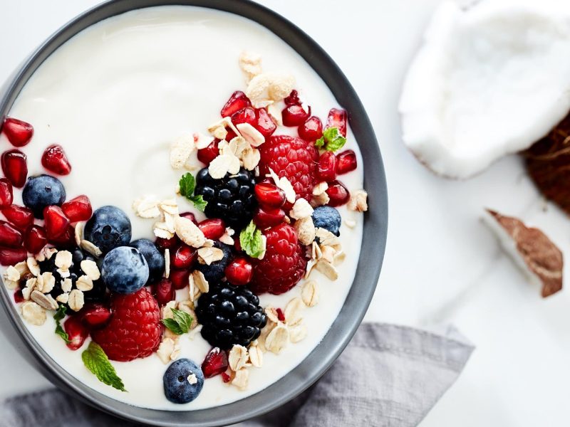 Draufsicht, heller Untergrund: graue Schüssel mit Kokos-Joghurt selber machen, darauf Beeren, Granatapfelkerne, Minze und Haferflocken, daneben Kokosnuss.