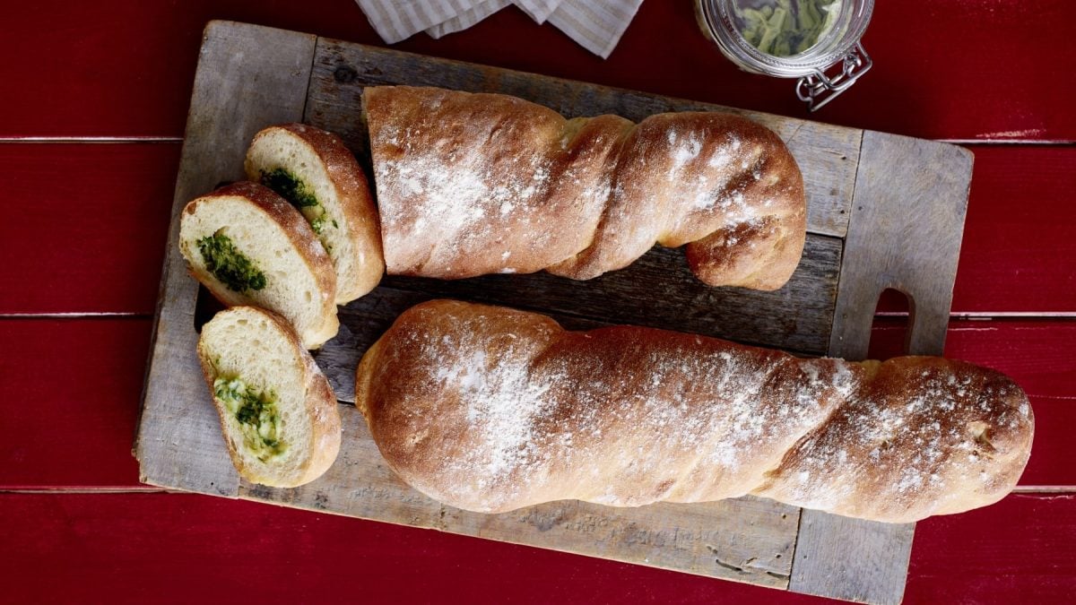 Angeschnittenes Kräuterbaguette auf einem Holzbrett auf rotem Holzuntergrund.