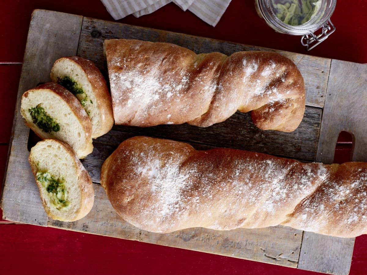 Angeschnittenes Kräuterbaguette auf einem Holzbrett auf rotem Holzuntergrund.