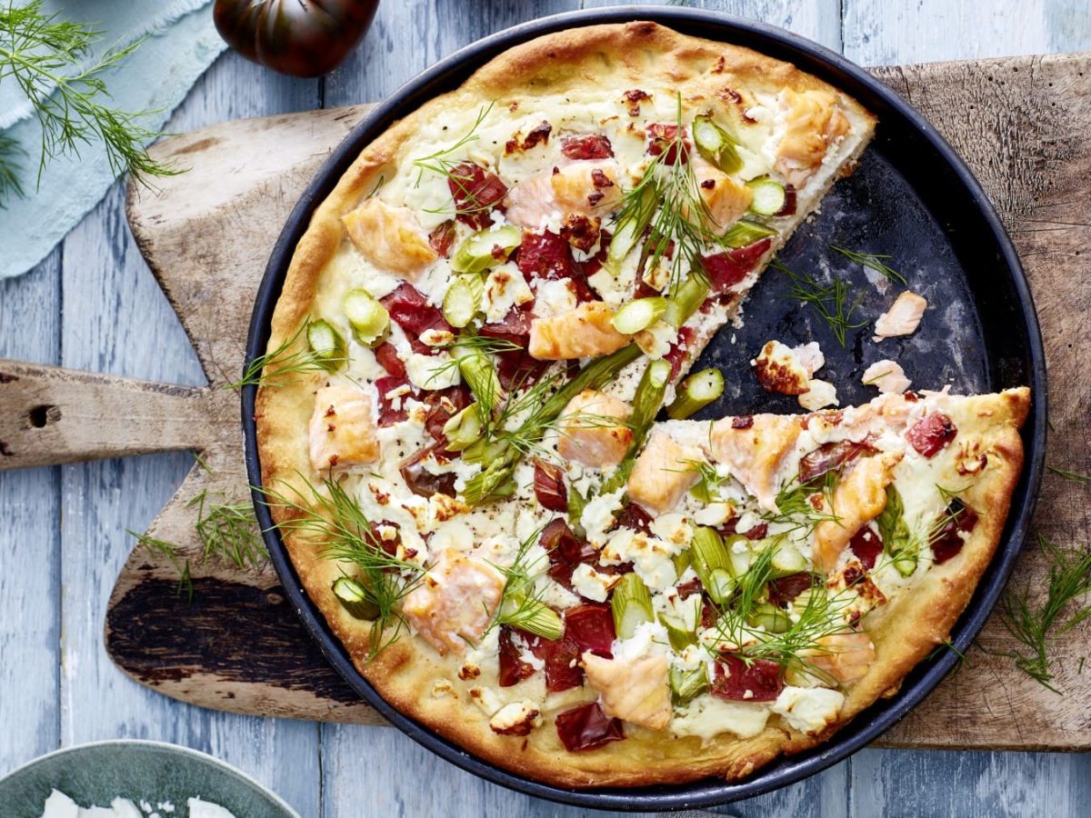 Eine Lachs-Feta-Pizza auf einer schwarzen Pizza-Backform auf einem hölzernen Schneidebrett auf einem graublau-hölzernen Hintergrund. Links oben Dill und eine Tomate auf einem hellblauen Geschirrtuch.