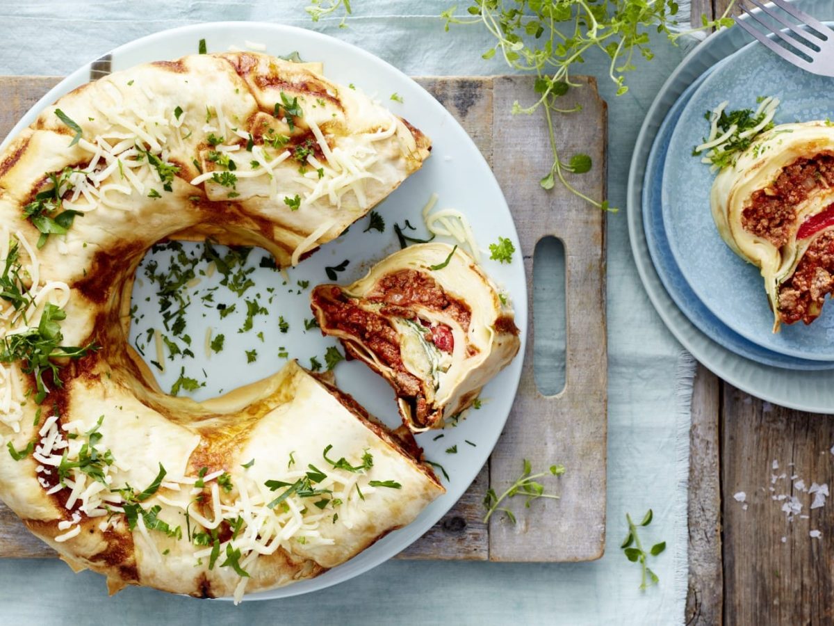 Ein weißer Teller mit einem angeschnittenen Lasagnering auf einem hölzernen Schneidebrett auf einem hellblauen Geschirrtuch auf einem hölzernen Hintergrund. Rechts ein blauer Teller mit einem Stück Lasagne, Kräuter und eine kleine Schüssel mit Salz. Links auf dem Bild drei Gabeln.