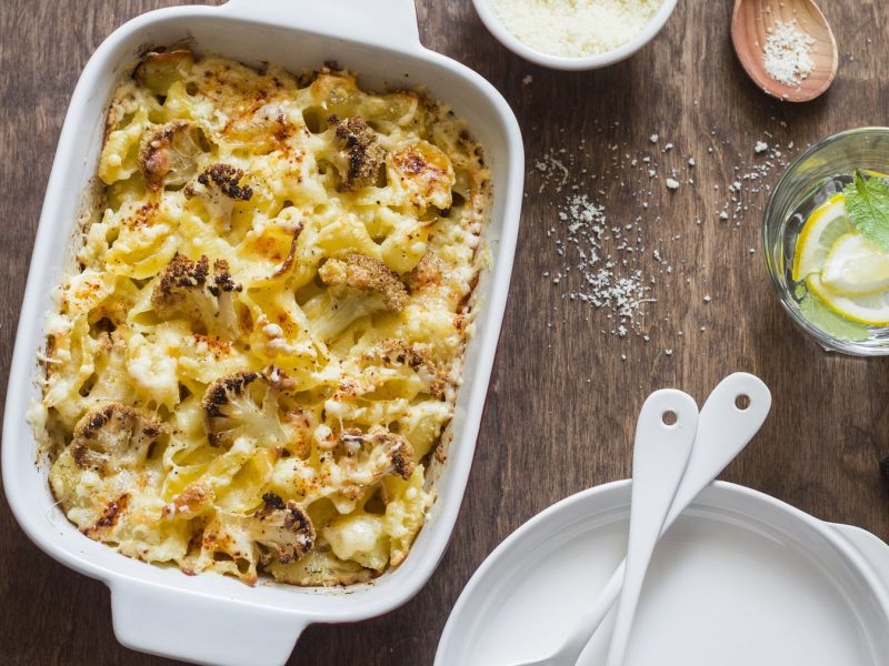 Weiße Auflaufform mit den Mac and cheese mit Blumenkohl auf einem Holztisch.