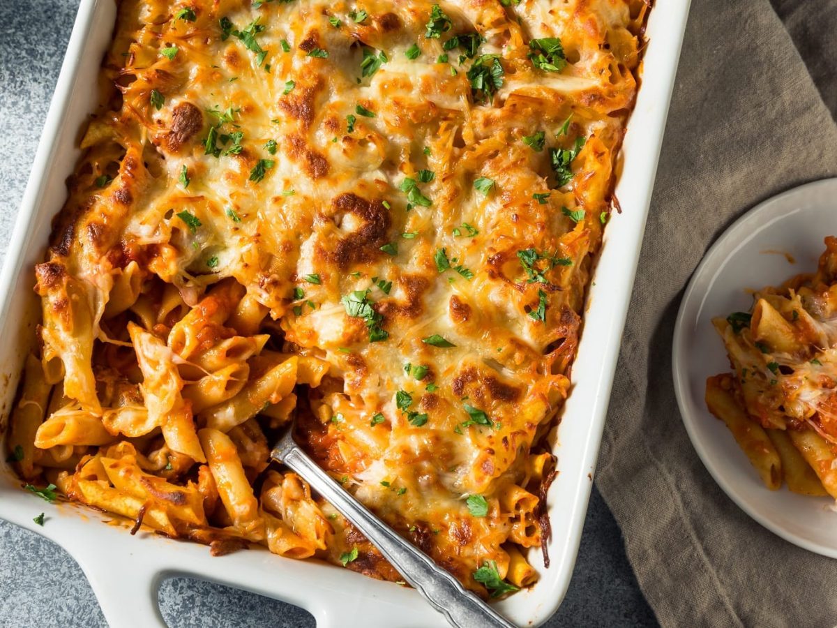 Eine weiße Auflaufform mit den überbackenen Mostaccioli-Nudeln auf einem grauen Tisch von oben fotografiert. Daneben eine kleine weiße Schale mit einem Stück vom Auflauf.