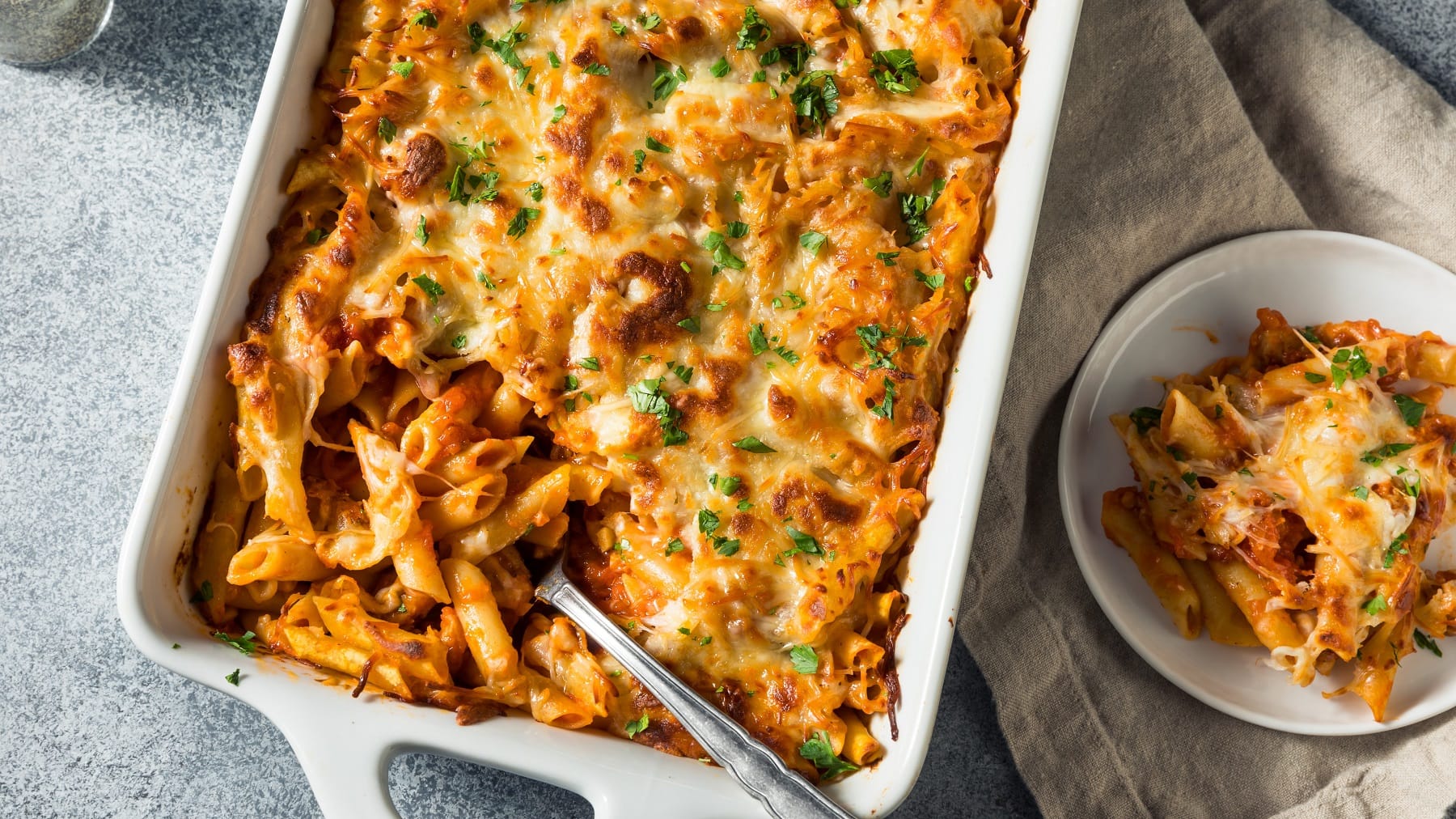 Eine weiße Auflaufform mit den überbackenen Mostaccioli-Nudeln auf einem grauen Tisch von oben fotografiert. Daneben eine kleine weiße Schale mit einem Stück vom Auflauf.