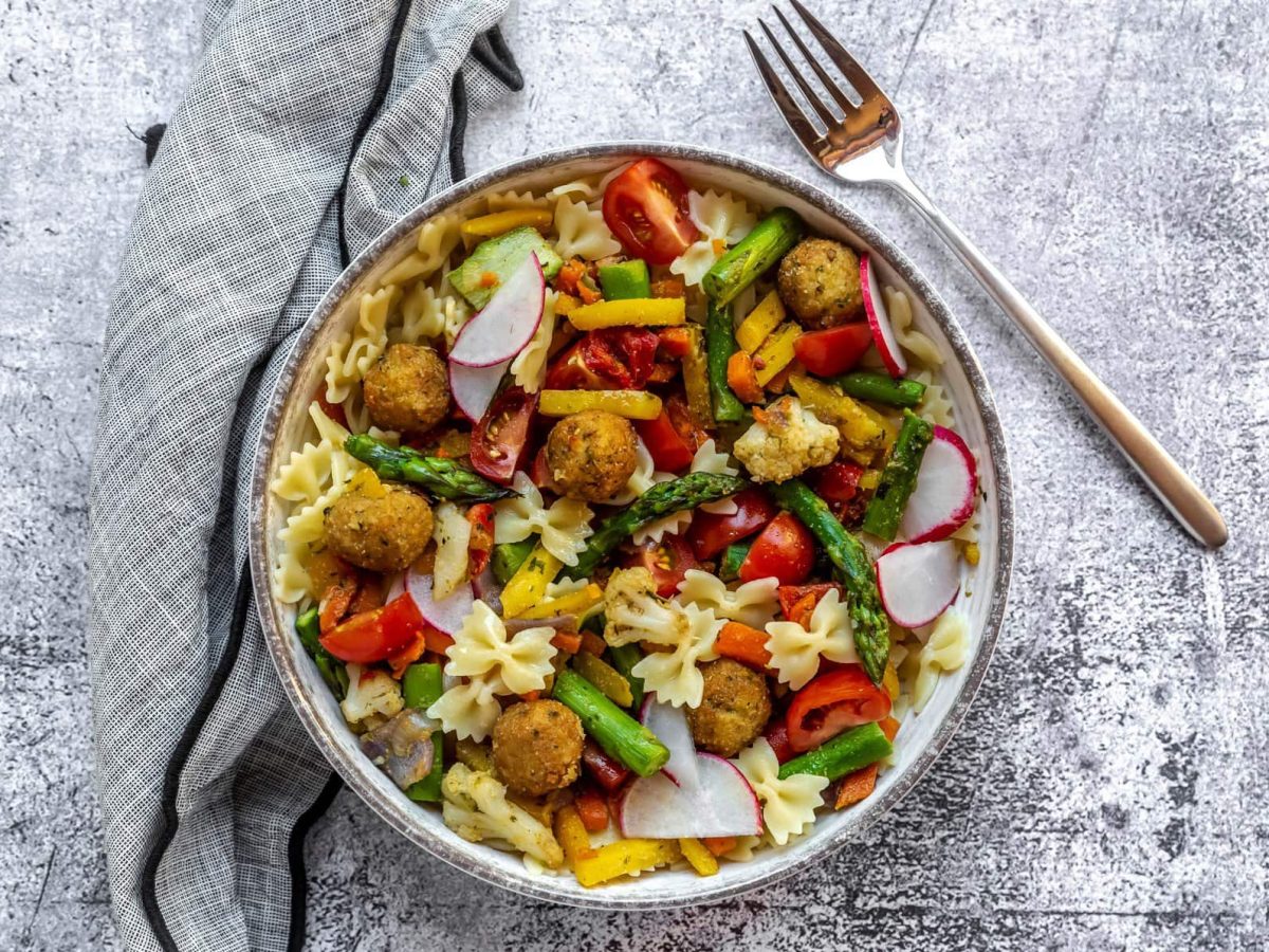 Nudelsalat mit grünem Spargel, Falafel und Gartengemüse in einem Teller auf grauem Untergrund. Im Bild eine Gabel. Draufsicht.