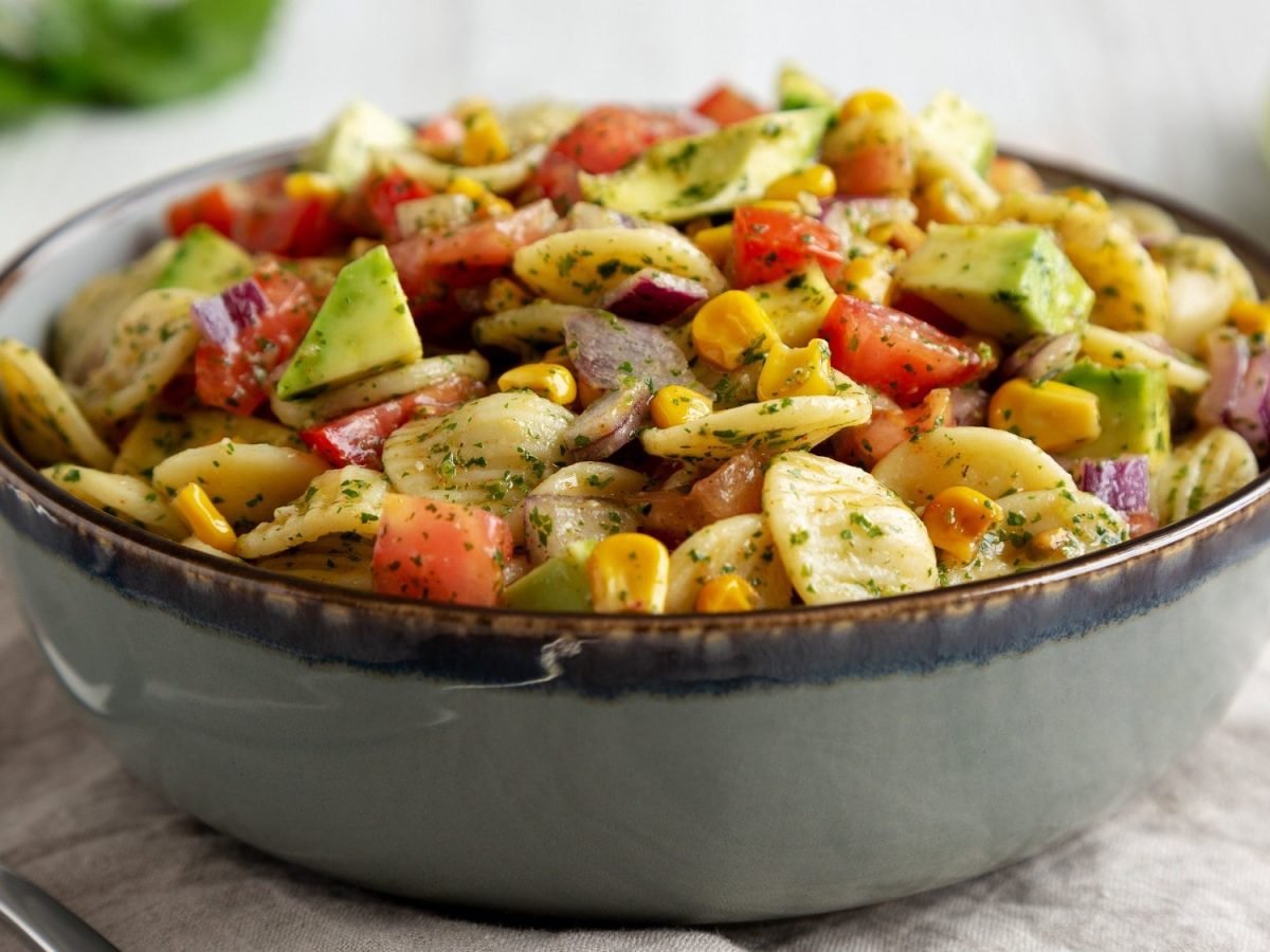 Eine Schüssel Nudelsalat mit Mais und Avocado sowie Paprika auf weißem Untergrund.