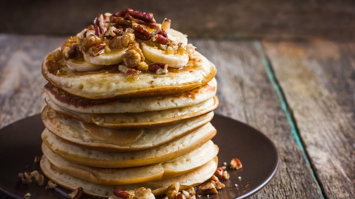 Mehrere Pancakes auf einem schwarzen Teller mit Erdnussbutter und Nüssen.