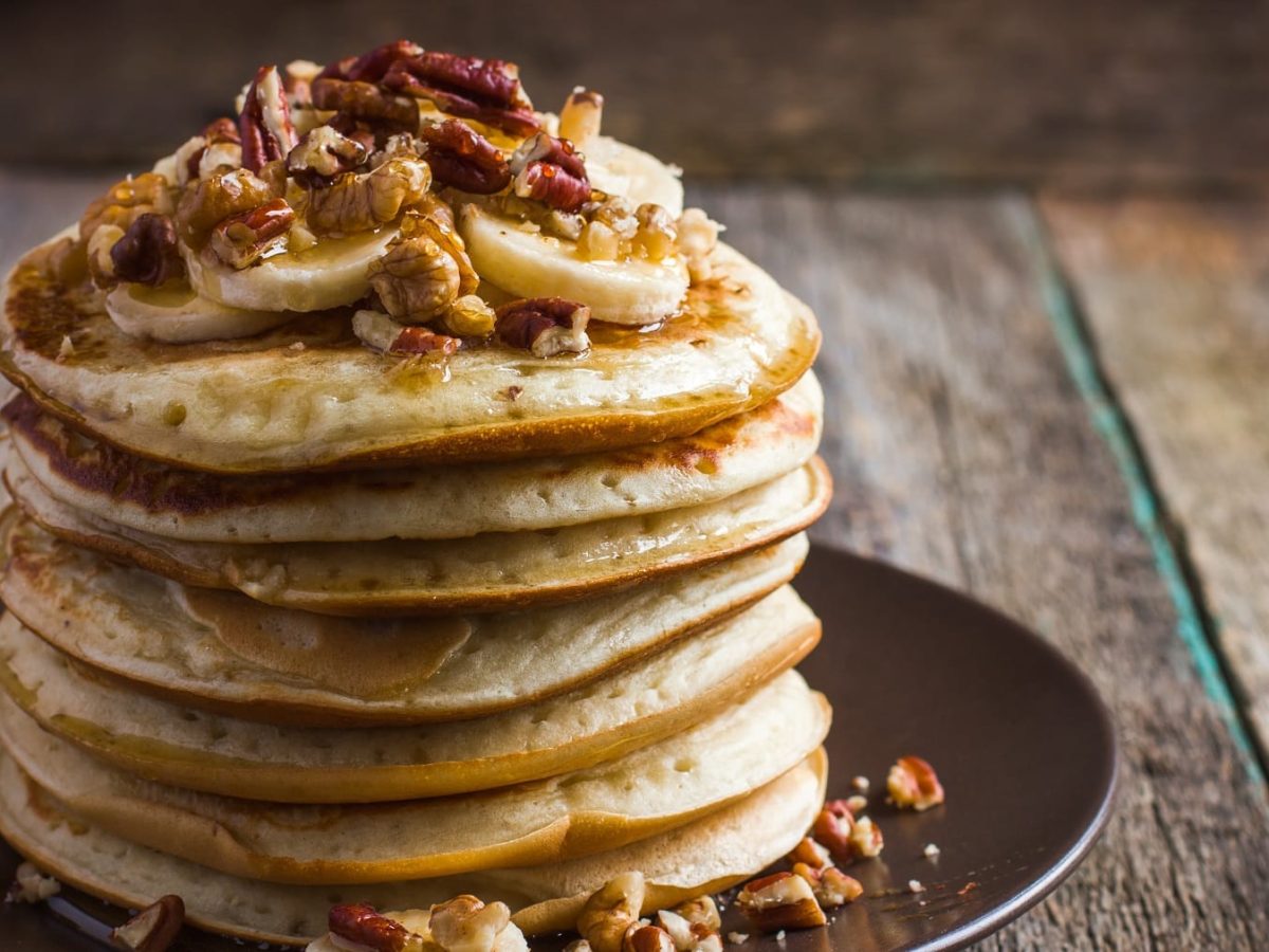 Mehrere Pancakes auf einem schwarzen Teller mit Erdnussbutter und Nüssen.