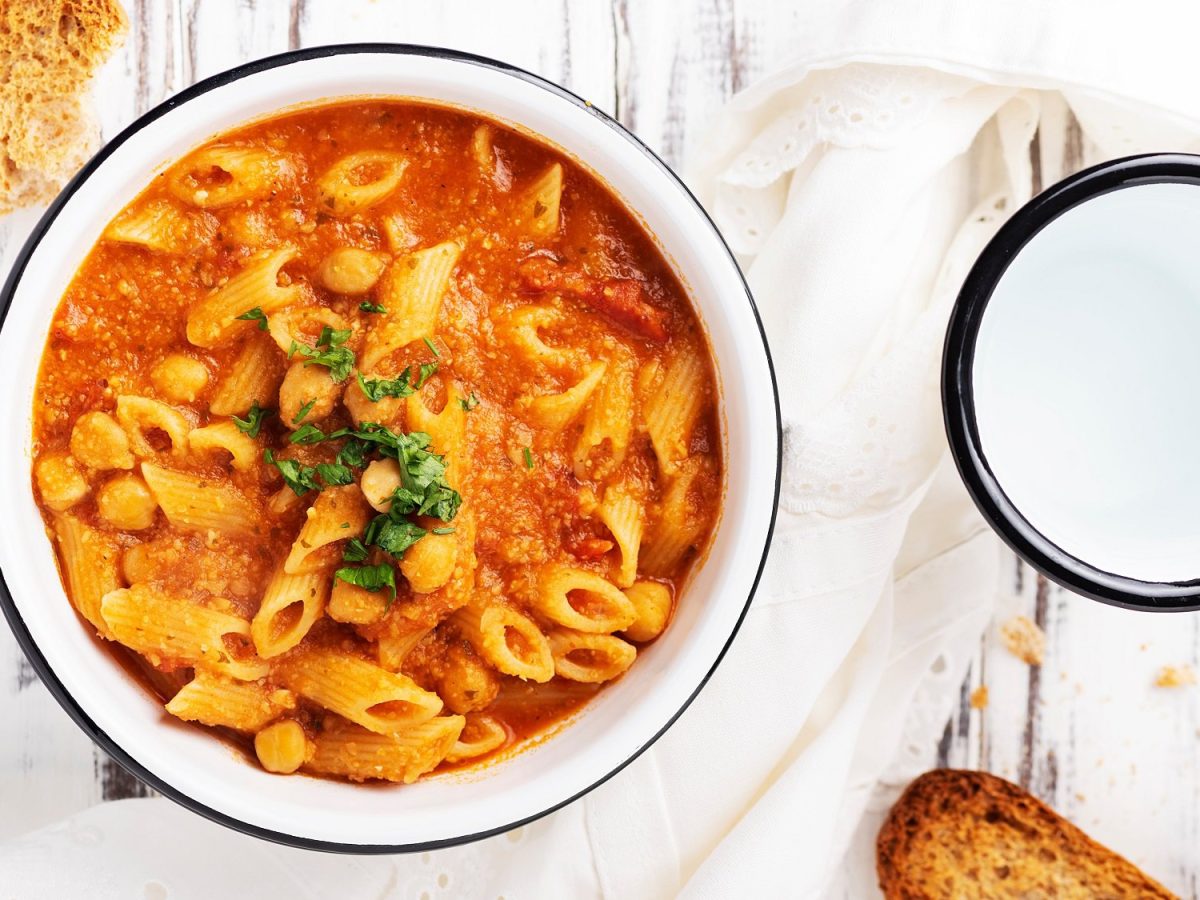 Eine Schüssel Pasta e ceci, ein italiensicher Nudeleintopf mit Kichererbsen und Tomatensuppe, in der Draufsicht, daneben ein Becher Wasser und geröstetes Brot.