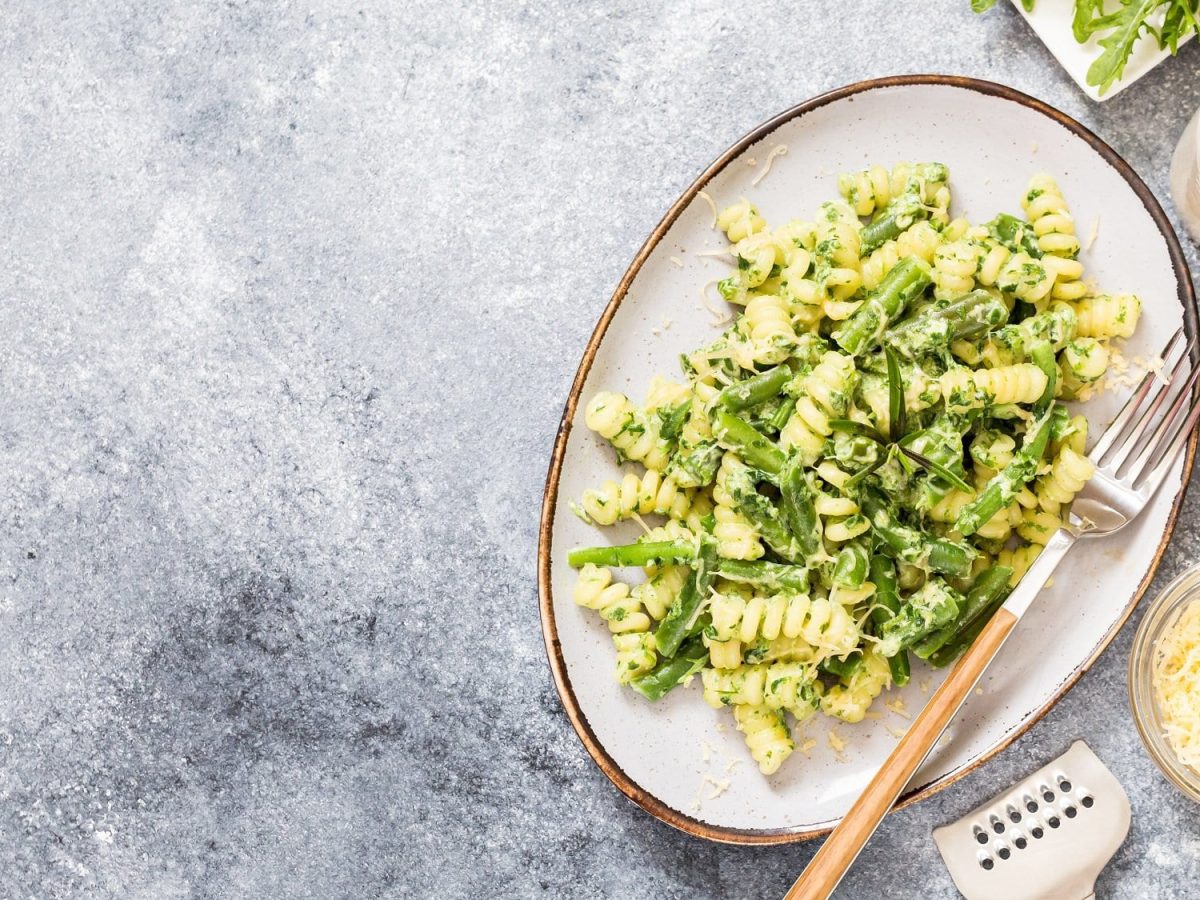 Ein Teller Pasta mit Spargel und Rucola, darauf auch eine Gabel, daneben Salz- und Pfefferstreuer, eine Schale mit geriebenem Käse, alles Draufsicht.