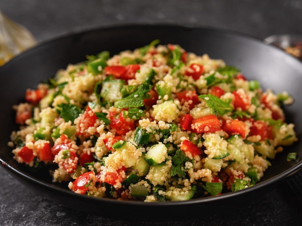 Ein Teller Pesto-Couscous-Salat mit Minze, Paprika, Gurke und Tomate, daneben eine Gabel.