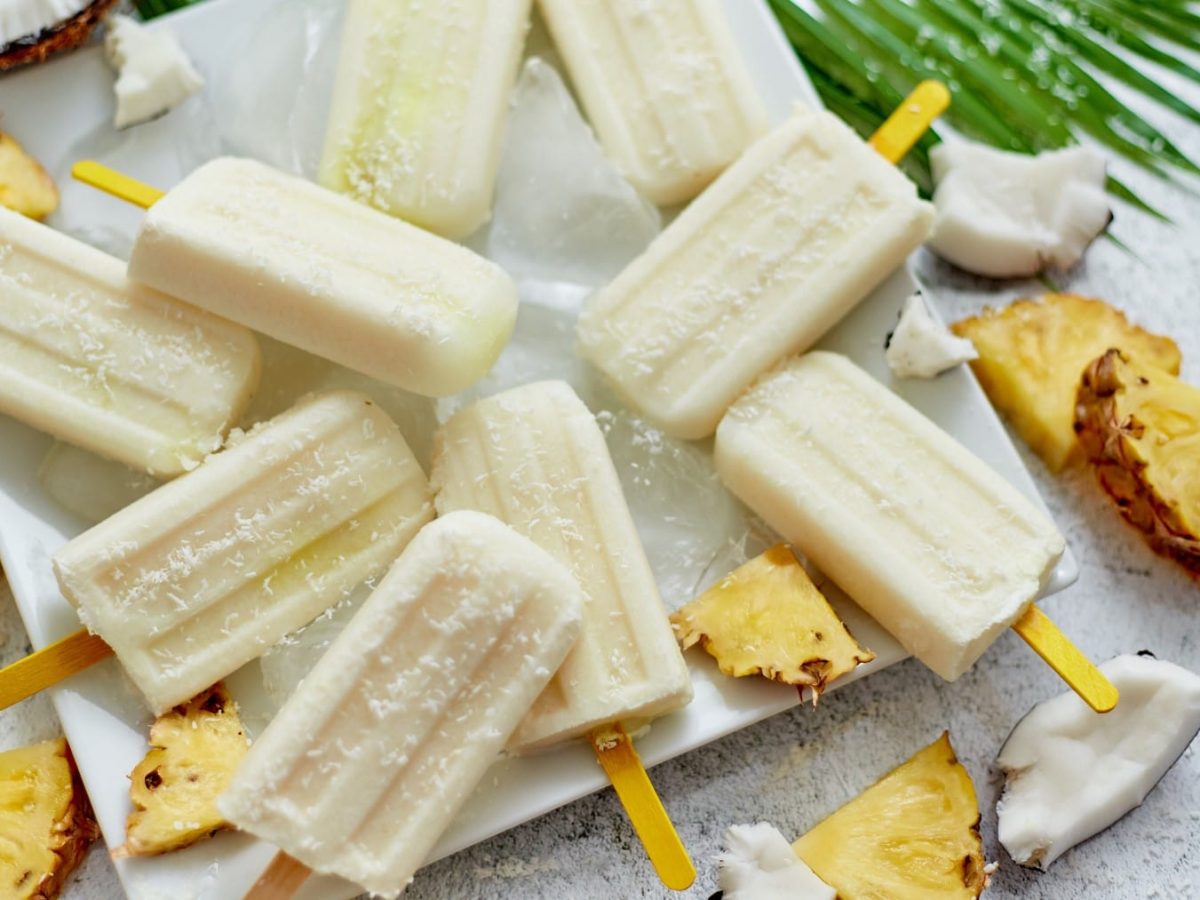 Mehrere Piña Colada Paletas auf einem weißen Teller. Drum herum Ananas- und Kokosstücke auf grauem untergrund.