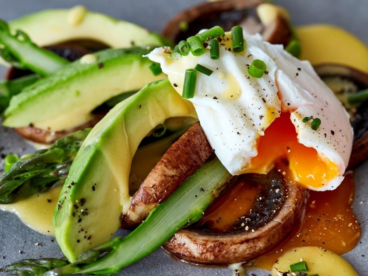 Nahaufnahme von Pilzen, grüner Spargel, pochiertes Ei, Avocado und Schnittlauch auf einem grauen Teller.