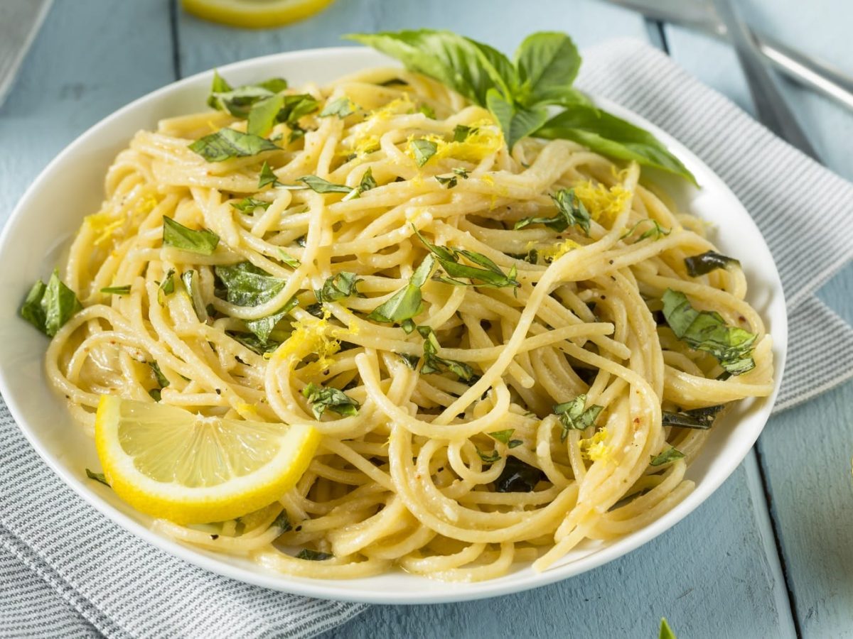 Ein weißer Teller mit der Ricotta Lemon Pasta auf einem hellen Holztisch mit grauen Tischdecken.